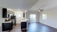 This single-floor space showcases a thoughtfully designed open-concept layout that seamlessly connects the kitchen and living area. Rich, dark hardwood floors run throughout, lending warmth and a contemporary feel to the interior. The kitchen is well-appointed with stainless steel appliances, including a refrigerator and gas range, complemented by sleek dark cabinetry and a peninsula with a built-in sink — ideal for both meal preparation and casual dining. Upper cabinet accents in matching dark tones add a cohesive, modern aesthetic. The living area is bright and airy, benefiting from multiple windows that invite natural light from multiple directions. A ceiling fan with a brushed metal finish provides both comfort and a stylish focal point overhead. Sliding glass doors open to a private balcony, offering a glimpse of lush greenery beyond. The neutral gray and white wall palette keeps the space feeling clean and versatile, while recessed lighting in the kitchen adds functional illumination. The overall ambiance is modern and move-in ready, with a clean architectural design that balances practicality with contemporary style.