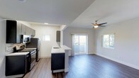 This single-floor space showcases a thoughtfully designed open-concept layout that seamlessly connects the kitchen and living area. Rich, dark hardwood floors run throughout, lending warmth and a contemporary feel to the interior. The kitchen is well-appointed with stainless steel appliances, including a refrigerator and gas range, complemented by sleek dark cabinetry and a peninsula with a built-in sink — ideal for both meal preparation and casual dining. Upper cabinet accents in matching dark tones add a cohesive, modern aesthetic. The living area is bright and airy, benefiting from multiple windows that invite natural light from multiple directions. A ceiling fan with a brushed metal finish provides both comfort and a stylish focal point overhead. Sliding glass doors open to a private balcony, offering a glimpse of lush greenery beyond. The neutral gray and white wall palette keeps the space feeling clean and versatile, while recessed lighting in the kitchen adds functional illumination. The overall ambiance is modern and move-in ready, with a clean architectural design that balances practicality with contemporary style.