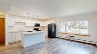 This well-appointed 2-bedroom, 1-bathroom single-floor unit offers a functional and modern layout across a comfortable footprint. The open-concept kitchen and living area serves as the heart of the home, anchored by a central island with a light countertop that provides additional prep space and casual seating. The kitchen is outfitted with crisp white shaker-style cabinetry, black appliances including a refrigerator, electric range, and microwave, and a stainless sink. Three pendant lights hang above the island, adding both task lighting and a contemporary design touch, while recessed lighting and a large window flood the space with natural light and outdoor views. Wood-look plank flooring extends throughout the kitchen and living areas, giving the space a clean, cohesive feel. The living room offers an open, versatile space with baseboard heating and an additional wall-mounted AC unit for year-round comfort. The primary bedroom features wood-look flooring and a walk-in closet with built-in shelving, while the secondary bedroom is carpeted and includes a window with neighborhood views and baseboard heating. The full bathroom includes a tub/shower combo, a pedestal sink, and a mirrored medicine cabinet.