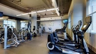 This fitness center offers a thoughtfully designed workout space spread across a single floor, providing ample room for a full range of exercise activities. The layout is open and organized, with dedicated zones for cardio equipment and strength training. A row of elliptical machines and steppers lines one side of the room, offering users a variety of low-impact cardio options, while additional machines such as cable systems and resistance equipment occupy the central and rear areas. The industrial-style ceiling features exposed ductwork, conduit, and bright linear fluorescent lighting, lending the space a modern, urban aesthetic. A wall-mounted television is positioned near the windows, providing entertainment during workouts. Natural light filters in through a large window along one wall, complementing the overhead lighting to keep the space bright and energizing. The flooring is durable rubber, ideal for high-traffic fitness use. Mirrored wall panels in one section of the room enhance the sense of space and allow users to monitor their form. A decorative artwork piece adds a touch of personality to the otherwise functional environment, contributing to a clean and motivating atmosphere.
