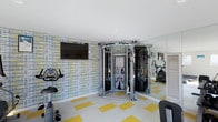 This single-floor fitness space is thoughtfully designed to accommodate a variety of workout routines within a compact footprint. The room features a bold, patterned accent wall in blue, yellow, and white tones, adding a vibrant energy to the space. The flooring complements this aesthetic with a checkerboard-style pattern of white and yellow tiles, creating a lively and motivating atmosphere.

At the center of the room stands a multi-functional cable machine, offering a wide range of strength training exercises. Cardio equipment, including a stationary bike, is positioned along the walls, maximizing usable floor space. A wall-mounted flat-screen television is installed on the accent wall, allowing users to stay entertained during workouts.

A full-length mirror spans one wall, visually expanding the room while serving a practical purpose for monitoring exercise form. Recessed ceiling lighting provides even, consistent illumination throughout. A louvered panel, likely concealing storage or utility access, is integrated along one side of the room, maintaining a clean and organized appearance.