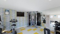 This single-floor fitness space is thoughtfully designed to accommodate a variety of workout routines within a compact footprint. The room features a bold, patterned accent wall in blue, yellow, and white tones, adding a vibrant energy to the space. The flooring complements this aesthetic with a checkerboard-style pattern of white and yellow tiles, creating a lively and motivating atmosphere.

At the center of the room stands a multi-functional cable machine, offering a wide range of strength training exercises. Cardio equipment, including a stationary bike, is positioned along the walls, maximizing usable floor space. A wall-mounted flat-screen television is installed on the accent wall, allowing users to stay entertained during workouts.

A full-length mirror spans one wall, visually expanding the room while serving a practical purpose for monitoring exercise form. Recessed ceiling lighting provides even, consistent illumination throughout. A louvered panel, likely concealing storage or utility access, is integrated along one side of the room, maintaining a clean and organized appearance.