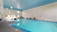 This single-level indoor pool facility is designed with both functionality and comfort in mind. The centerpiece is a generously sized rectangular swimming pool with clear, blue-tinted water, well-suited for lap swimming and recreational use. The pool deck surrounds the water on multiple sides, providing ample space for entry and exit, with a stainless steel ladder for easy pool access.

The interior is characterized by crisp white vertical paneling along the walls, which contributes to a clean, classic aesthetic. The ceiling is painted a soft sky blue, complementing the color of the pool water and enhancing the airy atmosphere of the space. Pendant globe lights hang from the ceiling, providing ambient illumination throughout.

Natural light filters in through a row of windows along one wall, brightening the space and offering a connection to the outdoors. Potted tropical plants are positioned around the pool deck, adding touches of greenery and a welcoming feel. A small seating area near the windows provides a comfortable spot for relaxation. Safety signage is posted on the walls, reflecting the facility's attentiveness to guest safety.