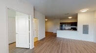 This single-floor unit is designed with a clean, contemporary aesthetic and an open-concept layout that connects the living area to the kitchen seamlessly. Wood-style laminate flooring runs throughout the main living spaces, providing a cohesive and low-maintenance foundation. The kitchen is well-appointed, featuring rich dark cabinetry along both walls, granite countertops, black appliances including a range, refrigerator, and over-the-range microwave, and a stainless steel sink. A raised peninsula with a granite surface extends into the living area, offering additional prep space and a natural division between the kitchen and living room without sacrificing openness. The living area is generously sized, with white walls and recessed flush-mount lighting providing a neutral, bright backdrop. A dedicated entry door opens directly into this main living space. The bedroom is a comfortable, well-proportioned room featuring a ceiling fan with light, a window with blinds admitting natural light and outdoor views, and a closet. The bathroom includes tile flooring, a full tub and shower combination, a toilet, and a vanity with a granite countertop and dark wood cabinetry. Throughout, white trim and panel-style doors maintain a consistent, polished finish.