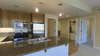 This single-floor residence offers a thoughtfully designed open layout centered around a stylish, functional kitchen. The kitchen is a standout feature, showcasing rich dark granite countertops with a double stainless steel sink set into a spacious island that doubles as a breakfast bar. Warm medium-toned wood cabinetry provides ample storage, complemented by stainless steel appliances including a refrigerator, range, and built-in microwave. Pendant lighting hangs above the island, adding both task illumination and a refined aesthetic touch. Recessed ceiling lights throughout ensure consistent, well-distributed lighting across the space. The open-concept design allows for a smooth visual flow from the kitchen into the adjacent living and hallway areas, where hardwood-style flooring continues the warm tone established by the cabinetry. A hallway visible from the kitchen leads to additional rooms, hinting at a practical and connected floor plan. Neutral wall tones and clean white trim work throughout contribute to a bright, inviting ambiance. The overall design balances functionality with contemporary style, making it a comfortable and efficient living environment.