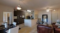 This single-floor residence showcases an open-concept layout that seamlessly connects the kitchen, dining, and living areas into one cohesive space. The kitchen is equipped with stainless steel appliances, including a refrigerator, and features a peninsula with dark granite-style countertops that doubles as a breakfast bar, providing both additional prep space and casual seating. White cabinetry lines the kitchen walls, complemented by open shelving displaying small decorative accents.

A striking dark brown accent wall anchors the dining area, creating a bold contrast against the otherwise neutral white walls and light-colored carpeted flooring throughout. A pendant light fixture hangs above the dining space, adding a warm focal point. The living area is generously proportioned, furnished with a leather sofa and a patterned accent chair, with framed artwork adorning the walls.

A ceiling fan is visible in the adjoining room, suggesting additional living or bedroom space beyond. The overall ambiance is warm and welcoming, combining functional design with tasteful decorative touches that create a comfortable, livable environment.