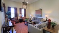 This single-story living space is designed with comfort and style in mind, immediately drawing attention with its notably high ceilings that lend an open, airy feel to the room. A striking wrought-iron lantern-style chandelier serves as a focal point overhead, adding an element of classic elegance to the space.

The living area is generously proportioned, accommodating multiple seating arrangements that create distinct yet cohesive conversation areas. A pair of patterned armchairs upholstered in rich jewel tones anchor one side of the room, complemented by boldly printed curtains in deep red and burgundy that frame the natural light streaming through multiple windows. On the opposite side, a neutral gray sectional sofa provides a modern contrast, accented by a large abstract canvas artwork mounted on the wall behind it.

A geometric-patterned area rug in red and cream ties the seating areas together, adding warmth and visual interest underfoot. Wooden accent tables, table lamps, and carefully placed decorative objects contribute to the layered, well-appointed aesthetic. Overall, the space balances traditional and contemporary elements to create an inviting and functional environment.
