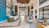 Located at 881 W Warren Ave in Longwood, Florida, this striking single-floor communal space spans a generous footprint and makes an immediate impression with its soaring double-height ceilings and open, airy layout. The grand entrance lobby is anchored by a sleek, marble-faced reception desk set against a dramatic teal accent wall adorned with large-scale heron artwork, creating a bold focal point upon arrival. Warm wood slat partitions and black-framed glass panels define zones throughout the space while maintaining a sense of visual openness.

The living area is thoughtfully composed, featuring an exposed brick accent wall rising to the full height of the ceiling, complemented by built-in shelving and curated wall art. A distinctive organic wood coffee table, leather and upholstered sofas, and layered area rugs add warmth and texture. Floor-to-ceiling windows dressed with elegant striped drapery flood the interior with natural light and offer views of lush Florida palms and outdoor amenities beyond.

Patterned tile inlays punctuate the large-format porcelain floors, while a statement chandelier of branching gold metalwork illuminates the double-volume ceiling above the reception area. Multiple seating vignettes throughout the space offer versatile areas for gathering, relaxing, or informal meetings, all unified by a cohesive coastal-modern design palette of teal, white, natural wood, and earthy brick tones.