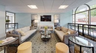 The community room is thoughtfully designed to accommodate both relaxed socializing and functional gatherings. The space is divided into two distinct yet complementary zones: a welcoming lounge area and a fully equipped kitchen.

The lounge area is furnished with plush neutral-toned sofas and accent chairs arranged around a circular glass coffee table, all anchored by a patterned area rug. Soft blue-grey walls, framed artwork, and a wall-mounted flat-screen television contribute to a polished, contemporary atmosphere. Floor-to-ceiling arched windows along one wall flood the seating area with natural light and offer views of the surrounding outdoor streetscape, lending an open, airy quality to the space.

The kitchen is outfitted with light wood cabinetry, dark countertops, and a white subway tile backsplash. Stainless steel appliances—including a refrigerator, double wall ovens, a cooktop with a suspended range hood, and a dishwasher—make the space well-suited for catering or communal cooking. A large island with bar-height seating for multiple guests serves as a central gathering point. Light wood-toned flooring ties both areas together, creating a cohesive and inviting environment throughout.