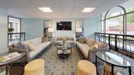 The community room is thoughtfully designed to accommodate both relaxed socializing and functional gatherings. The space is divided into two distinct yet complementary zones: a welcoming lounge area and a fully equipped kitchen.

The lounge area is furnished with plush neutral-toned sofas and accent chairs arranged around a circular glass coffee table, all anchored by a patterned area rug. Soft blue-grey walls, framed artwork, and a wall-mounted flat-screen television contribute to a polished, contemporary atmosphere. Floor-to-ceiling arched windows along one wall flood the seating area with natural light and offer views of the surrounding outdoor streetscape, lending an open, airy quality to the space.

The kitchen is outfitted with light wood cabinetry, dark countertops, and a white subway tile backsplash. Stainless steel appliances—including a refrigerator, double wall ovens, a cooktop with a suspended range hood, and a dishwasher—make the space well-suited for catering or communal cooking. A large island with bar-height seating for multiple guests serves as a central gathering point. Light wood-toned flooring ties both areas together, creating a cohesive and inviting environment throughout.