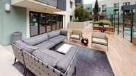 This contemporary outdoor amenity space is set within a multi-story residential community, thoughtfully designed to provide residents with a variety of functional and recreational areas. The courtyard is anchored by a generously sized outdoor lounge area, furnished with deep-seated sectional sofas, accent chairs, and a central coffee table arranged atop a patterned area rug laid over warm wood decking — creating an inviting setting for relaxation and socializing. Large sliding glass doors connect the outdoor deck to interior amenity spaces, allowing for a seamless indoor-outdoor flow. Adjacent to the lounge, an outdoor fitness zone features stationary cycling bikes positioned on the same wood deck, with a fully equipped fitness room visible through floor-to-ceiling glass behind them. A warm, horizontally clad cedar structure serves as a dedicated media area, complete with an embedded screen and posted usage guidelines, offering an open-air entertainment experience. Raised masonry planters border the courtyard walkways, filled with lush, layered landscaping including bamboo, philodendron, and ornamental grasses that add privacy and a natural aesthetic. Architectural elements throughout include modern tile and concrete finishes, ambient courtyard lighting, and patio heaters, all framed by the surrounding multi-story building facades.