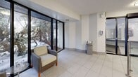 The lobby at 200 Ridley Blvd in Toronto offers a functional and well-organized entry space spread across a single floor. The interior is finished with large-format white ceramic tile flooring that runs consistently throughout, lending a clean and cohesive aesthetic to the space.

The reception and waiting area is furnished with modern upholstered seating, including armchairs positioned to take advantage of the floor-to-ceiling windows that frame views of mature trees and the surrounding greenery — providing a calm, natural backdrop to the interior. The two-tone wall treatment, featuring light grey wainscoting beneath white upper walls, adds a subtle layer of architectural detail throughout.

Circulation is well-served by multiple access points: a stainless steel elevator with tiled surrounds provides vertical access, while a stairwell with metal-edged steps connects to other levels of the building. Glass and stainless steel railings line a ramped entry corridor, offering accessible passage between entry points. Recessed and surface-mounted ceiling lighting ensures the space is evenly lit throughout.

The hallways connect the various functional zones of the lobby, including mailbox stations and intercom panels, supporting the day-to-day needs of residents. Overall, the lobby presents a practical, tidy, and accessible environment suited to a mid-rise residential building.
