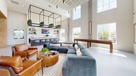 This single-floor amenity space is a thoughtfully designed communal lounge that seamlessly blends modern aesthetics with functional comfort. The defining architectural feature is the dramatic double-height ceiling, which floods the interior with natural light through a sweeping bank of tall windows, creating an open and airy atmosphere throughout.

The space is divided into two distinct zones. The lounge area anchors one end of the room with large-format gray porcelain tile flooring, a generous sectional sofa in rich blue velvet, leather accent chairs, and a round wood coffee table. Built-in light wood shelving units flank a wall-mounted television, providing display space and adding warmth to the clean, white walls. A wooden game table adds a recreational element near the windowed wall.

The kitchen and bar area occupies the opposite end, featuring a striking large island with a white quartz countertop, ornate wood-turned legs, and bar-height seating. The surrounding cabinetry is finished in warm natural wood tones, complemented by a white tile backsplash, open floating shelves, and a wall-mounted television. Cascading greenery suspended from a bold black metal overhead structure brings vibrant life and texture to the space, while pendant lighting with gold accents adds a refined finishing touch.