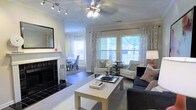 This single-floor residence offers a thoughtfully designed living space that balances comfort and style. The main living area serves as the heart of the home, featuring a striking fireplace with a black surround and a classic white mantle as a focal point. A ceiling fan with integrated lighting and a complementary track lighting system ensure the room is well-lit at any time of day. Multiple windows dressed with elegant patterned drapes flood the space with natural light, offering views of the surrounding greenery outside. The living area is furnished with a coordinated arrangement of dark upholstered sofas, white accent chairs, and a minimalist coffee table, creating a welcoming and functional layout. Crown molding along the ceiling adds a refined architectural touch throughout. Adjacent to the living room, a separate dining area is visible, featuring additional seating and continued access to natural light. The consistent use of hardwood flooring, neutral wall tones, and carefully selected décor elements contributes to a cohesive and inviting overall ambiance, making this space both practical and aesthetically pleasing.