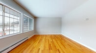 This single-floor space presents a straightforward and functional layout connecting several well-defined rooms via a central hallway. The corridor, finished with warm honey-toned hardwood floors, leads to multiple doorways, providing clear separation between living areas. The main living space is generously proportioned, featuring the same rich hardwood flooring and neutral gray-toned walls. Large multi-pane windows allow natural light to fill the room, with views of the surrounding outdoor landscape, including trees and open surroundings. Baseboard heating units run along the perimeter walls throughout the unit, providing consistent warmth. The bedrooms are carpeted in a neutral beige tone, maintaining a comfortable and quiet feel. Each bedroom includes a large window framing outdoor views and a baseboard heating unit. The kitchen is equipped with warm oak cabinetry extending both above and below the countertops, offering ample storage. White appliances — including a refrigerator, electric range, and range hood — accompany a double stainless steel sink. The floor is finished in neutral tile. The full bathroom includes a tiled tub-shower combination, a vanity with oak cabinetry, tile flooring, and a baseboard heater. Overall, the space maintains a clean, neutral palette throughout.