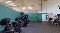This single-level recreational facility is thoughtfully designed to support a wide range of fitness and leisure activities. The fitness area is divided into dedicated zones, each serving a distinct purpose. The cardio section features a row of SportsArt ellipticals and upright bikes lined along teal-accented walls, with rubber flooring for safety and comfort. Large industrial fans provide airflow, and a wall-mounted television keeps users entertained during workouts. A strength training zone offers a cable machine, rowing machine, medicine balls, and various resistance equipment, all reflected in full-wall mirrors that amplify the sense of space. Overhead, recessed skylights and flush panel lighting illuminate the gym with a bright, energizing atmosphere. Adjacent to the fitness area, an indoor basketball court provides a spacious, open floor with regulation-style markings and a wall-mounted adjustable hoop. Soaring ceilings and clerestory windows allow natural light to filter in, creating a clean and airy environment ideal for active play. A separate game room rounds out the facility, featuring carpeted flooring, high ceilings, natural light from upper windows, and recreational staples including a foosball table and ping pong table — perfect for casual socializing and friendly competition.