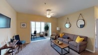 Located at 1047 Hwy 31 in Spanish Fort, Alabama, this single-floor residence offers a comfortable and thoughtfully arranged living environment. The main living area is defined by warm yellow walls that create an inviting, cheerful atmosphere, complemented by soft carpeted flooring that adds to the overall coziness of the space. A ceiling fan overhead provides practical comfort while contributing to the room's understated charm. The living area is generously sized, accommodating a full seating arrangement centered around a rustic wooden coffee table, offering ample room for relaxation and entertaining. Natural light flows freely into the space through a series of windows visible in the adjoining sunlit nook or sitting area, which features large windows that bathe the room in bright, airy light. This secondary area provides an ideal spot for reading or casual seating and is separated from the main living space by a wide, open doorway that maintains a connected, open-concept feel throughout. Decorative touches such as wall art, a round hanging mirror, and accent pillows lend personality and a well-curated aesthetic to the interior. The overall ambiance is warm, bright, and livable, making this a welcoming single-floor home in the Spanish Fort community.