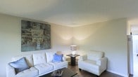 This single-floor residential space offers a thoughtfully designed interior with a contemporary aesthetic and a calm, neutral color palette. The living area serves as the focal point of the home, featuring white tufted upholstered seating, including a sofa and matching armchair, arranged around a dark wood coffee table. A bold abstract canvas painting in muted earth tones anchors one wall, adding an artistic element to the space. A stylish table lamp with a sculptural base provides warm accent lighting, complementing the ambient illumination that casts a soft glow across the walls. The flooring consists of dark hardwood, which provides a rich contrast to the light-colored furniture and walls. The ceiling features a textured finish, adding subtle visual interest overhead. The overall ambiance is clean, uncluttered, and inviting, with a modern sensibility that balances simplicity with carefully chosen decorative touches. The neutral tones and cohesive design elements throughout the living area create a welcoming atmosphere well-suited for both relaxing and entertaining.