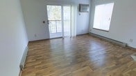 This two-floor space offers a clean, functional layout designed to maximize comfort and utility. The main living area is finished with warm, rich hardwood-style flooring that runs throughout, lending a cohesive and inviting atmosphere to the space. Large windows allow generous amounts of natural light to fill the room, creating a bright and airy environment throughout the day. A sliding glass door opens directly onto a private balcony, providing outdoor access and pleasant views of the surrounding greenery — a notable feature that adds to the overall appeal of the space. Vertical blinds offer privacy and light control at the sliding door. A wall-mounted air conditioning unit ensures year-round climate comfort, complemented by a baseboard heating system along the exterior wall. The neutral white walls provide a versatile backdrop suitable for a variety of interior styles. The overall ambiance is open, clean, and well-lit, with practical features thoughtfully integrated throughout. Spanning two floors, this space offers ample room for a range of uses, with each level contributing to the sense of spaciousness and livability that defines the property.