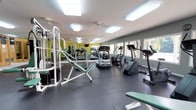 This fitness center is designed to support a well-rounded exercise routine in a clean, functional environment. The space features durable rubber flooring throughout, providing safety and comfort during workouts. Cardio equipment includes multiple treadmills and an elliptical machine, strategically positioned to allow ample space between each unit. A multi-station cable and weight machine anchors the strength training area, accompanied by adjustable workout benches for a variety of exercises. Large mirrors line one wall, enhancing the sense of openness while allowing users to monitor their form. The walls are painted in a fresh combination of white and sage green, contributing to an energizing yet calm atmosphere. Bright flush-mount ceiling fixtures ensure the space is well-lit throughout the day, while a row of windows along one side invites natural light and provides a connection to the outdoors. A ceiling fan adds ventilation and comfort during intense workouts. The layout is thoughtfully organized to maximize usability, offering sufficient open floor space for stretching or bodyweight exercises. Overall, the fitness center delivers a practical and welcoming setting for users of all fitness levels.