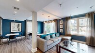 Located at 5680 Bay St in Emeryville, California, this thoughtfully designed community lounge offers a versatile and inviting space across a single floor. The interior is defined by bold teal and deep blue accent walls that lend a contemporary, energetic character throughout, complemented by warm wood-tone flooring that runs seamlessly from room to room.

The lounge area features a generously sized sectional sofa with teal throw pillows, anchored beneath a striking sputnik-style chandelier with globe bulbs. Large windows dressed in sheer linen curtains flood the space with natural light and offer views of the surrounding community outdoors. A media wall with a large flat-screen television and a stylish credenza provides an entertainment focal point.

The dining area accommodates multiple white-topped tables paired with modern black chairs, making it well-suited for collaborative gatherings or casual meals. Framed artwork adorns the teal walls, adding a curated aesthetic to the setting.

The full kitchen is a standout feature, showcasing rich wood-grain cabinetry, white quartz countertops, a geometric-patterned tile backsplash, stainless steel appliances including a side-by-side refrigerator and dishwasher, and a large central island with pendant lighting overhead. The space balances functionality with refined design, making it an ideal amenity for residents.
