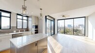 Located in Houston's desirable 77027 zip code, this thoughtfully designed single-floor residence offers three bedrooms and three bathrooms across a well-appointed layout. The open-concept kitchen and living area serves as the heart of the home, showcasing light hardwood flooring, floor-to-ceiling sliding glass doors, and sweeping Houston skyline views. The chef's kitchen is outfitted with stainless steel appliances, a gas range, grey shaker cabinetry, a subway tile backsplash, and a substantial center island topped with granite countertops, illuminated by globe pendant lights. The living area extends toward a wall of glass, with a ceiling fan and abundant natural light creating a bright, airy atmosphere. The primary bedroom features plush carpet, black-framed windows with city views, and direct access to a well-appointed en-suite bathroom complete with a granite vanity, wood-look tile flooring, and a frameless glass walk-in shower. Additional bathrooms mirror the same refined finishes, including a soaking tub with tiled surround. Bedrooms are serviced by spacious closets with built-in shelving, and an in-unit washer and dryer adds everyday convenience. The hallway connects all rooms seamlessly, maintaining a clean, modern aesthetic throughout.