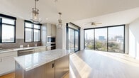 Located in Houston's desirable 77027 zip code, this thoughtfully designed single-floor residence offers three bedrooms and three bathrooms across a well-appointed layout. The open-concept kitchen and living area serves as the heart of the home, showcasing light hardwood flooring, floor-to-ceiling sliding glass doors, and sweeping Houston skyline views. The chef's kitchen is outfitted with stainless steel appliances, a gas range, grey shaker cabinetry, a subway tile backsplash, and a substantial center island topped with granite countertops, illuminated by globe pendant lights. The living area extends toward a wall of glass, with a ceiling fan and abundant natural light creating a bright, airy atmosphere. The primary bedroom features plush carpet, black-framed windows with city views, and direct access to a well-appointed en-suite bathroom complete with a granite vanity, wood-look tile flooring, and a frameless glass walk-in shower. Additional bathrooms mirror the same refined finishes, including a soaking tub with tiled surround. Bedrooms are serviced by spacious closets with built-in shelving, and an in-unit washer and dryer adds everyday convenience. The hallway connects all rooms seamlessly, maintaining a clean, modern aesthetic throughout.