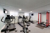 This fitness center at 3009 W 13 Mile Rd in Royal Oak, Michigan, offers a thoroughly equipped workout facility designed to support a full range of exercise routines. The space features durable interlocking rubber tile flooring throughout, providing both safety and comfort during workouts. The drop-panel ceiling with recessed lighting ensures the room is consistently and evenly illuminated.

The cardio section is well-stocked with multiple treadmills, elliptical trainers, and upright stationary bikes, several of which feature integrated digital displays. A wall-mounted television is positioned for convenient viewing during cardio sessions. A drinking fountain is conveniently installed along one wall.

The strength training area is equally impressive, featuring a bold red power rack with a flat bench, a multi-functional cable machine with an adjustable bench, and a fully stocked three-tier dumbbell rack. Bosu balance trainers and additional benches round out the functional training options. A large wall mirror on one side of the room visually expands the space and aids in form monitoring during exercises.

The walls are finished in a combination of crisp white and a calm light teal accent, contributing to a clean and energizing atmosphere. The layout is thoughtfully organized to allow ample circulation between equipment zones, making efficient use of the available floor space.