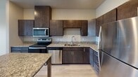 Located at 1408 21st Ave in Austin, Minnesota, this single-floor unit offers a thoughtfully designed layout with modern finishes throughout. The kitchen is a standout feature, outfitted with rich dark-stained cabinetry that extends to the ceiling for ample storage, granite countertops, and a full suite of stainless steel appliances including a range, microwave, dishwasher, and refrigerator. A kitchen island with a matching granite surface provides additional workspace and casual dining potential. Light hardwood-style flooring runs through the main living areas, lending a clean and contemporary feel. The open layout connects the kitchen to the living area, which benefits from natural light and a wall-mounted climate control unit. The bedroom is carpeted for comfort and features a window with outdoor views and a baseboard heating unit. The bathroom is finished with a granite-topped dark wood vanity, a framed mirror, and grab bars, indicating accessibility-conscious design. Light wood-tone flooring carries through the bathroom as well, maintaining a cohesive aesthetic. Overall, the unit presents a functional and well-appointed interior suited for comfortable single-level living.