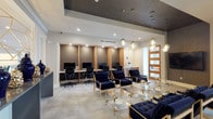 This polished, single-floor commercial space showcases a refined modern aesthetic defined by a cohesive palette of navy, gold, and warm wood tones. The central lobby and lounge area serves as the heart of the space, furnished with deep navy tufted velvet seating arranged around sleek acrylic-and-gold coffee tables. Overhead, a dramatic coffered ceiling accent features a cascading cluster of globe pendant lights, creating an eye-catching focal point that elevates the overall ambiance. Large-panel wood-accented doors with frosted glass inserts frame the perimeter, allowing natural light to filter through while maintaining privacy. A front reception counter provides a welcoming point of arrival for guests. Adjacent to the lounge, a dedicated business center offers multiple individual workstations, each housed within wood-paneled alcoves equipped with desktop computers and ergonomic chairs — ideal for productivity in a professional setting. A glass-enclosed conference room is accessible via a sliding glass partition and a solid wood door, outfitted with a round table, white chairs, and warm pendant lighting, offering a private meeting environment. The flooring transitions between large-format tile in the lounge and soft carpet in the office areas. Recessed lighting throughout ensures the entire space is well-illuminated, contributing to its professional yet inviting character.
