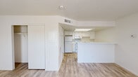 This single-floor unit offers a clean, minimalist aesthetic defined by crisp white walls, light wood-style flooring, and an abundance of natural light throughout. The open-concept living area greets you upon entry through a front door flanked by two large windows fitted with vertical blinds, allowing sunlight to pour across the smooth flooring and create an airy, welcoming atmosphere.

The kitchen is thoughtfully designed in a galley-style layout and is accessible via a peninsula that separates it from the living space. White cabinetry lines both sides of the kitchen, providing generous storage, while a skylight panel overhead brightens the workspace. The kitchen is equipped with a coil-top electric range, an over-the-range microwave, a refrigerator, and ample counter space on both sides.

The bedroom is a well-proportioned, serene space bathed in soft light, featuring the same wood-style flooring and a large walk-in closet with a rod and open shelving—offering practical and accessible storage. The bathroom presents a clean, functional design with a wide vanity, an integrated sink, and a large wall mirror that reflects light throughout the room. A separate closet or storage area is also visible nearby.

Overall, the unit maintains a consistent, bright palette and a straightforward, functional layout well-suited for comfortable everyday living.