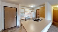 This single-floor residence offers a practical and comfortable layout designed for everyday living. The open-concept living and dining area is finished with wall-to-wall carpeting and features track lighting overhead, a wall-mounted air conditioning unit, and a sliding glass door that opens to a private balcony with outdoor views of surrounding greenery. The kitchen is positioned adjacent to the living space and is defined by a peninsula countertop with a double-basin sink, white cabinetry with warm wood-tone trim, a white electric range, refrigerator, and microwave. Tile flooring in the kitchen and entry areas provides a clean contrast to the carpeted living spaces. The bedrooms are generously sized, each featuring carpet flooring, baseboard heating, and ample natural light from large windows. Closet space is well-provided, with at least one bedroom offering a wide reach-in closet with a hanging rod. Two bathrooms serve the unit — one featuring a full tub/shower combo with a white surround, a wood-tone vanity, and tile flooring, and a second half-style bathroom with a vanity and tile floor. A dedicated in-unit laundry room houses a full-size washer and dryer, wire shelving, and an electrical panel, rounding out the functional amenities of this well-organized residence.