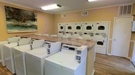 This communal laundry facility is thoughtfully designed to serve residents with efficiency and comfort in mind. The room features a generous lineup of top-load washing machines positioned along the center of the space, complemented by a row of stacked dryer units lining the back wall, providing ample capacity for simultaneous use. A long granite-style folding counter runs centrally through the room, offering a practical surface for sorting and folding laundry. The warm yellow-toned walls create a welcoming atmosphere, while crown molding along the ceiling adds a subtle touch of refinement. Overhead fluorescent lighting ensures the space is bright and well-lit throughout. Wood-look flooring runs underfoot, lending warmth and easy maintainability to the room. Each dryer unit is clearly numbered for easy identification, reflecting an organized and user-friendly approach to shared amenity design. A decorative piece of wall art adds a homey aesthetic touch, elevating the space beyond purely utilitarian function. Ventilation is well-addressed with ceiling vents distributed throughout, ensuring a comfortable environment during use. Overall, this laundry facility combines practicality with a clean, pleasant aesthetic.