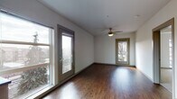 Located at 949 NE Orenco Station Loop in Hillsboro, Oregon, this single-floor unit is situated within the walkable Orenco Station community. The living area is finished with rich, dark hardwood-style flooring and features a ceiling fan with light fixture. Multiple windows and two glass-paned doors flood the space with natural light while providing direct access to a balcony, offering views of the surrounding neighborhood's mature trees and streetscape.

The kitchen is well-appointed with warm wood cabinetry extending to the upper walls, dark countertops, a herringbone-patterned tile backsplash, and a full suite of stainless steel appliances including a refrigerator, dishwasher, electric range, and built-in microwave. Flush-mount ceiling lights provide consistent illumination throughout the kitchen and adjoining areas.

The bedroom is carpeted and features a ceiling fan, a tray ceiling detail, and a large window with views toward the Orenco Station streetscape. The bathroom includes a wood-toned vanity with white countertop, a large mirror, and a full tub-shower combination with chrome fixtures. A dedicated in-unit laundry closet accommodates a stacked washer and dryer, adding everyday convenience to this thoughtfully designed space.