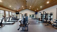 Located at 988 Old Shirley Rd in Central, SC, this single-floor fitness center offers a thoughtfully designed workout environment across a generously sized open layout. The main exercise area is defined by a striking vaulted ceiling clad in warm natural wood planking, fitted with recessed lighting that illuminates the entire space evenly. Large gridded windows line the walls, flooding the room with abundant natural light and providing views of an outdoor pool area, creating an energizing atmosphere for workouts.

The main gym floor is finished with durable rubber flooring and is outfitted with a comprehensive selection of TRUE Fitness commercial-grade equipment, including multiple treadmills, a stair climber, and an upright cycle for cardio training. The strength zone features cable machines, a leg curl/extension bench, a power rack with barbell and weight plates, and a dedicated dumbbell rack. Wall-mounted mirrors expand the sense of space and allow for proper form monitoring during strength training. Mounted flat-screen televisions are positioned throughout for entertainment.

A separate flex room provides a distinct space for functional fitness and group-style training. This room features vibrant patterned flooring, resistance bands, stability and medicine balls, foam rollers, exercise mats, and a striking full-height living green plant wall that adds a refreshing, natural element to the workout environment.