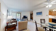 This 1-bedroom, 1-bathroom residence is thoughtfully laid out across a single floor, offering a comfortable and functional living environment. The combined living and dining area forms the heart of the home, featuring neutral-toned carpet, crisp white walls, and warm wood-trimmed accents throughout. A sliding glass door opens to a private balcony, inviting natural light and outdoor views of lush greenery into the space. The living area provides ample room for seating arrangements and entertainment, flowing seamlessly into the dining space. The kitchen is efficiently designed with warm honey-toned cabinetry, light countertops, tile flooring, and a full suite of appliances including a refrigerator, range, built-in microwave, and dishwasher. The bedroom is a quiet retreat featuring plush carpeting, a corner window with blinds for natural light and privacy, and a wide sliding-door closet system with built-in shelving for generous storage. The bathroom includes a warm wood vanity with an integrated sink, a large mirror with overhead lighting, and a shower-tub combination. A dedicated entry closet and additional storage are conveniently located near the foyer, which is accented by a pendant light fixture. Warm wood door accents are carried consistently throughout, creating a cohesive and inviting aesthetic.