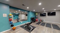 Located in Wyndamere, this fitness center is a thoughtfully designed, single-floor workout facility built to accommodate a wide variety of exercise styles and fitness goals. The space is finished with light wood-look luxury vinyl plank flooring throughout, providing a clean and durable surface ideal for both equipment use and floor exercises. Vibrant teal accent walls contrast with neutral gray walls and ceilings, creating an energizing yet balanced atmosphere. Recessed lighting is installed throughout the ceiling, ensuring the entire space is well-lit. The cardio zone features a treadmill, elliptical trainer, and an additional cross-trainer, all positioned on protective rubber mats. A dedicated strength area includes a multi-tier dumbbell rack with a full range of free weights, a weight bench, and a selection of medicine balls displayed on a wall-mounted rack. The open floor area accommodates multiple yoga mats laid out for group or individual stretching and mat-based workouts. A large wall mirror expands the sense of space and allows for form-checking during exercise. Two wall-mounted flat-screen televisions provide entertainment during workouts, and a decorative wall clock and small desk area add functional finishing touches to the room.