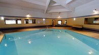 This indoor pool facility offers a welcoming aquatic environment designed for year-round use. The centerpiece of the space is a generously sized swimming pool with clear, light-turquoise water, illuminated by underwater lighting that gives the pool a bright and inviting appearance. The pool features a set of entry steps, making it easily accessible for swimmers of all ages and abilities.

The room is topped with an exposed beam ceiling finished in white, lending a classic, structured aesthetic to the space. Warm overhead lighting is mounted throughout, casting a comfortable glow across the water and surrounding areas. The walls are painted in a warm yellow tone and accented with a contrasting dark horizontal band, adding a defined sense of character to the interior.

Large mirrors are mounted along the walls, visually expanding the space and reflecting both the pool and the room's architectural details. A wooden bench is positioned along one wall, providing a convenient resting area for guests. Informational and safety signage is also displayed, indicating that the facility is well-maintained and regulation-compliant. A window allows a glimpse of natural light and greenery from outside, softening the enclosed atmosphere. Overall, the space presents a clean, functional, and comfortable environment for aquatic leisure.