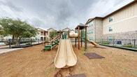 Located at 1215 E Owassa Rd in Edinburg, Texas, this outdoor playground serves as a dedicated recreational amenity within a residential community. The play area is fully enclosed by a sturdy black metal fence, providing a secure environment for children. The ground is covered with wood chip surfacing, offering a cushioned, safety-conscious base throughout the space.

At the heart of the playground stands a multi-level play structure finished in green and brown tones, featuring two tower platforms topped with green shingled roofs. The structure is equipped with multiple slides — including both straight and curved designs — as well as staircases, ramps, and climbing elements that encourage active play for a range of ages. Additional ground-level play equipment, including a spinning/rocking ride-on toy, is positioned in the open areas surrounding the main structure.

The playground is framed by mature shade trees that provide natural relief from the Texas sun. The surrounding multi-story residential buildings, constructed with a combination of stone veneer and board-and-batten siding, create a well-defined neighborhood backdrop. A gated entry point allows for controlled access, and the generous open footprint of the space provides ample room for free play beyond the structured equipment.