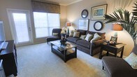 This single-floor space presents a thoughtfully designed living room that balances comfort and style. The room is anchored by a plush, dark charcoal sofa adorned with an assortment of patterned throw pillows, complemented by a coordinating upholstered armchair that invites relaxation. A sleek, dark-framed glass coffee table serves as the centerpiece, providing both function and visual interest.

The flooring features wall-to-wall carpeting in a warm neutral tone, contributing to the room's inviting ambiance. A large window dressed with a decorative fabric valance and horizontal blinds allows generous natural light to filter into the space, enhancing its open feel. Crown molding along the ceiling adds a refined architectural detail that elevates the overall aesthetic.

Wall décor includes a pair of circular mirrors and framed artwork, creating a curated gallery-like arrangement. A warm-toned table lamp provides soft accent lighting, while a potted plant introduces a subtle natural element. The cohesive color palette of browns, tans, and creams throughout gives the space a harmonious, polished appearance.