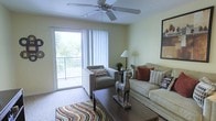 This single-floor residence offers a welcoming and well-appointed living space designed for both comfort and functionality. The living room serves as the heart of the home, featuring warm, neutral-toned walls that create a calm and inviting atmosphere. A ceiling fan with a light fixture is centrally positioned overhead, ensuring year-round comfort and air circulation throughout the room. Soft carpeting underfoot adds warmth and texture, complemented by a colorful area rug that anchors the seating arrangement. Large sliding glass doors with vertical blinds flood the space with abundant natural light and provide direct access to a private balcony, where lush greenery is visible, bringing a refreshing outdoor connection to the interior. The living area comfortably accommodates a full sofa and armchair arrangement centered around a dark wood coffee table, offering ample space for relaxing or entertaining. Decorative accents, including framed wall art and tasteful accessories, add personality to the room. The overall ambiance is bright, airy, and thoughtfully arranged, making this a practical and pleasant living environment.