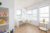 This single-floor studio is designed for efficient, comfortable living within a compact, open footprint. The layout integrates the living and sleeping areas seamlessly, with light wood-style flooring running throughout, contributing to a clean and cohesive aesthetic. Neutral white walls keep the space feeling bright and airy.

The kitchenette runs along one wall near the entry and includes a stainless steel sink, smooth-top cooktop, under-counter refrigerator, microwave, and open shelving above the continuous countertop — providing practical storage and prep space without occupying excessive room. A dedicated workspace with a desk and shelving unit is positioned near the windows, offering a functional area for productivity.

The living and sleeping zone benefits from multiple windows that allow generous natural light to fill the room, creating an open and welcoming atmosphere. Built-in open shelving provides additional organizational flexibility within the main living area.

The bathroom features tile flooring, a wall-mounted sink, toilet, and a towel bar, maintaining the unit's clean, minimal design language. Overall, the space is thoughtfully arranged to maximize functionality while maintaining a modern, uncluttered feel throughout.