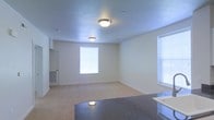 This single-floor space is designed with an open-concept layout that seamlessly connects the kitchen and living areas, creating a functional and cohesive environment. The living area features wall-to-wall carpeting in a neutral tone, contributing to a clean and understated aesthetic. Two windows fitted with white blinds allow natural light to fill the room, brightening the cream-colored walls and adding an airy quality to the interior.

Overhead, flush-mount ceiling light fixtures provide consistent ambient lighting across the space. The ceiling features a subtle texture and is finished in a light tone that complements the overall color palette.

The kitchen area is visible in the foreground, highlighted by a dark granite-style countertop that provides a contrasting accent against the lighter surroundings. A single-basin sink is positioned near the window, taking advantage of natural light. A door near the kitchen leads to an adjacent room, adding to the functional flow of the layout.

A floor-level wall vent is also present, indicating a built-in HVAC system. Overall, the space presents a straightforward, move-in-ready interior with practical finishes and a bright, open atmosphere.