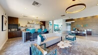 Located at The Grove in Phoenix, Arizona, this single-floor community clubroom offers a well-appointed gathering space designed with a bold, contemporary aesthetic. The open-plan layout seamlessly connects a comfortable lounge area with a fully equipped kitchenette, creating a cohesive and functional environment for residents to socialize and unwind.

The lounge area is anchored by a curved white sofa, patterned accent chairs in teal and navy, and a sculptural white coffee table, all set atop a textured, earth-toned carpet. A wall-mounted flat-screen television is paired with a sleek walnut media console, while a circular pendant light overhead adds a warm, modern focal point. Large windows allow natural light to flow in, offering views of the surrounding desert landscape, including mature palm trees.

The kitchenette features rich, dark walnut cabinetry, open floating shelves with decorative accents, a stainless steel refrigerator, and a generous white quartz island with vibrant turquoise bar stools beneath a striking gold starburst chandelier. A coffee station and full countertop workspace complete the functional amenity.

Glass-panel doors provide a visual connection to an adjacent fitness center, reinforcing the space's role as a central community hub. Colorful artwork and curated décor throughout contribute to the lively, resort-inspired atmosphere of this Phoenix amenity space.