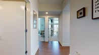 This well-appointed multi-bedroom unit offers a thoughtfully designed single-floor layout ideal for shared student living. A central hallway with rich hardwood flooring connects the various rooms, providing a clean and organized flow throughout the space. The open kitchen is equipped with stainless steel appliances, including a gas range, microwave, and refrigerator, complemented by sleek grey cabinetry and a peninsula countertop that separates the cooking area from the living space. The living room is bright and airy, benefiting from floor-to-ceiling windows that frame sweeping city skyline views and allow abundant natural light to fill the space. Multiple bedrooms are carpeted for comfort and each features large windows with urban vistas, built-in or freestanding wardrobes, a desk and chair for studying, and ample storage. One bedroom is configured as a double-occupancy room with two beds and two wardrobes. The unit includes two bathrooms: one full bath with a double-sink vanity, large mirror, tile flooring, and shower, and a second more compact bathroom also finished with tile floors and a shower. The overall aesthetic is modern and neutral, with white walls and clean lines throughout.