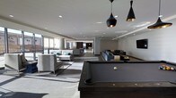 This single-floor amenity space offers a sophisticated, open-plan layout designed for social gathering and relaxation. The interior is anchored by a dark-finished billiards table positioned beneath a pair of striking black-and-gold pendant lights, which serve as a bold focal point against the bright white ceiling. Recessed lighting throughout ensures the entire space is evenly and elegantly illuminated. Multiple distinct seating areas are arranged across the open floor, each furnished with modern gray sofas and sectionals accented by teal throw pillows, lending subtle pops of color to the otherwise neutral palette. The light gray tile flooring transitions seamlessly across the space, reinforcing the cohesive, contemporary aesthetic. Large windows along the perimeter invite natural light deep into the interior, enhancing the bright and airy ambiance. Toward the rear, the space continues into additional lounge and social areas, hinting at the generous scale of the overall floor plan. The overall design sensibility is clean and minimal, with a refined industrial edge that creates an inviting yet upscale environment suited for both casual socializing and organized recreation.