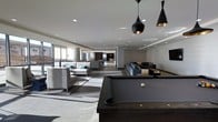 This single-floor amenity space offers a sophisticated, open-plan layout designed for social gathering and relaxation. The interior is anchored by a dark-finished billiards table positioned beneath a pair of striking black-and-gold pendant lights, which serve as a bold focal point against the bright white ceiling. Recessed lighting throughout ensures the entire space is evenly and elegantly illuminated. Multiple distinct seating areas are arranged across the open floor, each furnished with modern gray sofas and sectionals accented by teal throw pillows, lending subtle pops of color to the otherwise neutral palette. The light gray tile flooring transitions seamlessly across the space, reinforcing the cohesive, contemporary aesthetic. Large windows along the perimeter invite natural light deep into the interior, enhancing the bright and airy ambiance. Toward the rear, the space continues into additional lounge and social areas, hinting at the generous scale of the overall floor plan. The overall design sensibility is clean and minimal, with a refined industrial edge that creates an inviting yet upscale environment suited for both casual socializing and organized recreation.