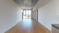 This single-floor space showcases a contemporary industrial aesthetic, blending raw architectural elements with modern finishes throughout. The open living area is defined by warm light hardwood flooring and an exposed concrete ceiling that lends an urban loft character to the space. Floor-to-ceiling windows flood the living area with abundant natural light while framing sweeping city views, creating a bright and airy atmosphere.

The kitchen is thoughtfully designed with high-gloss white cabinetry complemented by warm wood open shelving, a dark backsplash, and quartz-style countertops. A full suite of stainless steel appliances—including a refrigerator, range, built-in microwave, and dishwasher—provides a sleek, functional cooking environment. Recessed lighting adds to the polished finish.

The bedroom features plush blue carpeting, an exposed concrete column that reinforces the industrial character, and a tall vertical window offering additional city views and natural light. The bathroom is finished with a teal vinyl floor, white subway-tiled tub surround, a floating vanity with a neutral countertop, and clean white walls, delivering a crisp, modern feel throughout.