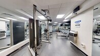 Located at 675 Richmond St in London, Ontario, this fitness center offers a thoughtfully organized exercise space across a single floor. The room is divided into distinct functional zones, maximizing the use of the available area. A dedicated cardio section houses treadmills and a rowing machine, positioned alongside a large wall mirror that enhances the sense of space and allows users to monitor their form. An adjacent spin cycle area features multiple stationary bikes equipped with individual screens, set against a bold, motivational typographic mural in vibrant colors — featuring words like "Dedication," "Push," and "Be Brave" — creating an energizing atmosphere. A wall-mounted flat-screen television accompanies this section. The strength training zone is equipped with a cable machine, a dumbbell rack, and a weight bench, catering to a variety of resistance exercises. The flooring transitions between durable rubber matting in the cardio and spin areas and polished concrete in the free weights zone. Overhead, a drop ceiling with flush-panel LED lighting provides bright, even illumination throughout. A bulletin board displaying workout references and a storage bench near the entrance add practical touches. The overall design is clean, functional, and motivating.