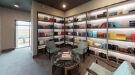 This thoughtfully designed space presents a refined blend of function and aesthetics across its single floor, showcasing a dramatic double-height interior visible through full-length glass partitions. The library lounge serves as the centerpiece, featuring wrap-around, floor-to-ceiling backlit bookshelves filled with a curated collection of books and decorative objects. The shelving is arranged in an L-shaped configuration in one area and in structured grid-style units in another, both bathed in warm recessed lighting that highlights the colorful arrangement of titles and accents.

Seating throughout is carefully selected, with patterned accent chairs, wingback chairs, and ottomans positioned to encourage reading and conversation. A round glass-top coffee table with a brass base anchors one of the sitting areas. The flooring transitions between soft carpet in the lounge zones and hard tile in the connecting corridors, adding subtle visual definition between spaces.

The corridor areas display large-scale abstract art pieces in bold teal, gold, and black tones, lending an elevated gallery-like quality to the transitional spaces. Glass walls and railings enhance openness and allow sightlines across levels. Large windows along the perimeter invite natural light and offer views to the surrounding exterior, further enriching the space's bright, contemporary ambiance.
